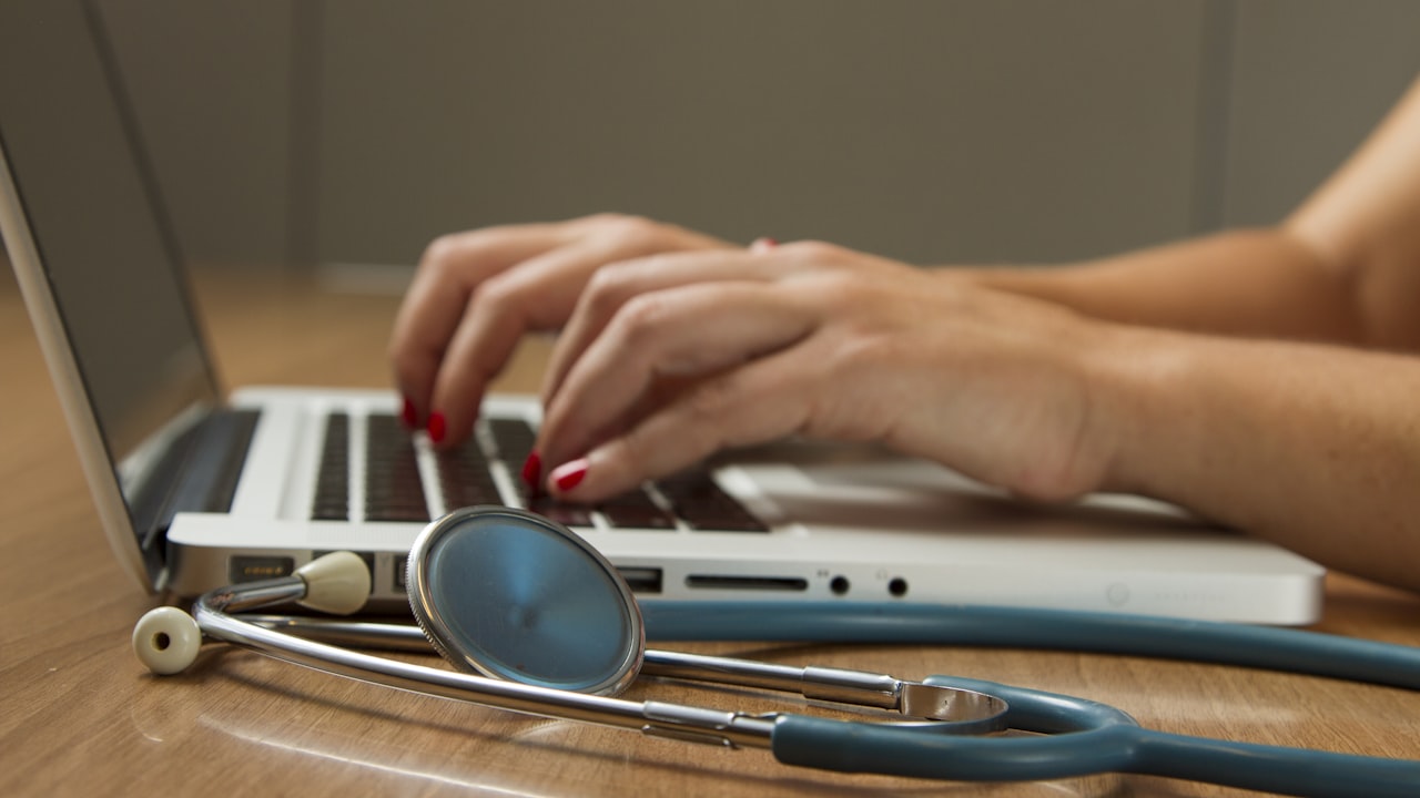 Healthcare professional typing on a laptop with a stethoscope on the desk